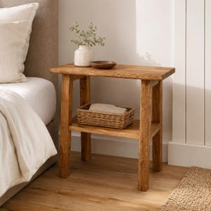 Puede incluir: Una mesa auxiliar rectangular de madera con acabado de veta natural, que cuenta con cuatro patas resistentes y un estante inferior. Un jarrón de cerámica blanca con una pequeña planta verde y un cuenco de madera marrón descansan sobre ella.