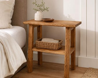 May include: A rectangular wooden side table with a natural grain finish, featuring four sturdy legs and a lower shelf. A white ceramic vase with a small green plant and a small brown wooden bowl sit on top, while a woven basket holds a folded cloth.