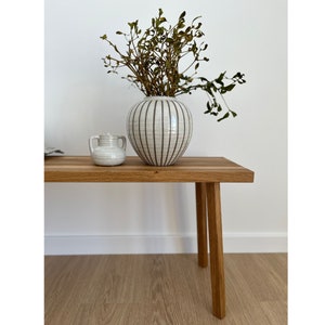May include: A wooden bench with four legs supports a white ceramic vase with black vertical stripes and a small white ceramic jar. The vase holds dried branches. The bench is made of light-colored wood.