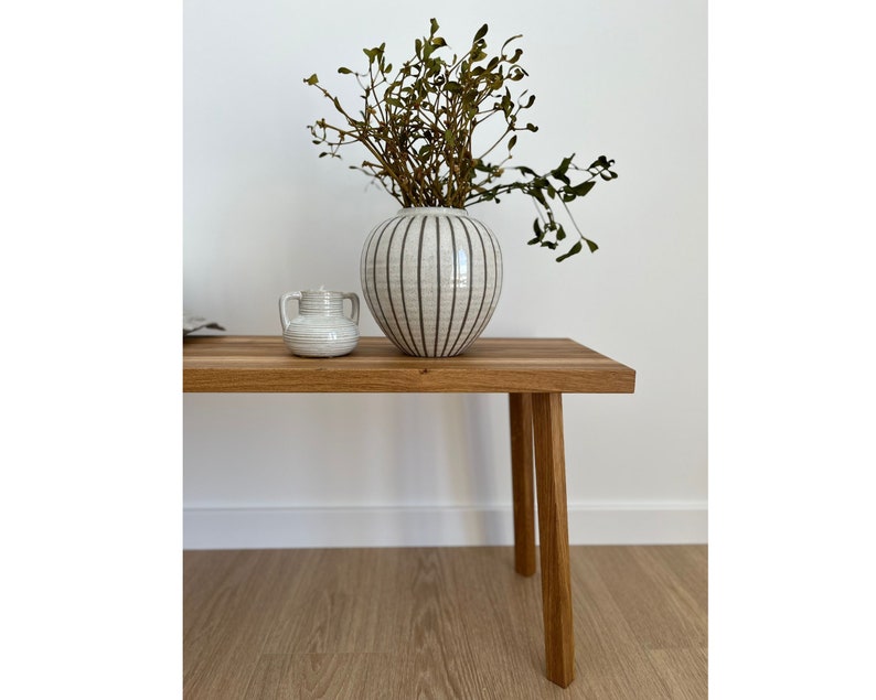 May include: A wooden bench with four legs supports a white ceramic vase with black vertical stripes and a small white ceramic jar. The vase holds dried branches. The bench is made of light-colored wood.