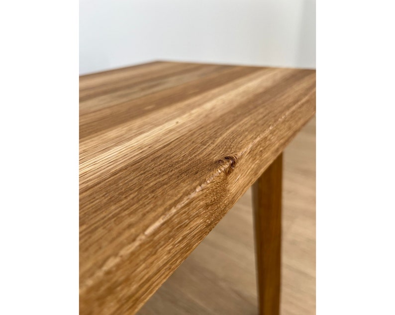 May include: Close-up of a wooden table with a natural wood grain finish. The table has a light brown color and a rectangular top. The legs are a similar color and are slightly tapered. The table is likely for indoor use.