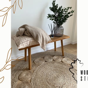 Puede incluir: Un banco de madera con una manta de color beige y una almohada con estampado rojo y blanco. El banco está sobre una alfombra de fibra natural con un patrón circular. Un jarrón con ramas verdes frondosas está sobre el banco.