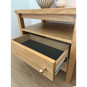 May include: A light oak wood nightstand featuring an open shelf and a single pull-out drawer. The drawer is equipped with a round, polished gold-tone metal knob and a black interior base. The drawer slides on visible metal side rails.