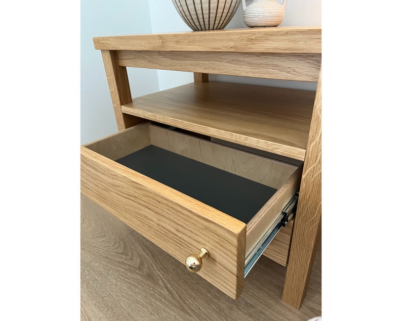 May include: A light oak wood nightstand featuring an open shelf and a single pull-out drawer. The drawer is equipped with a round, polished gold-tone metal knob and a black interior base. The drawer slides on visible metal side rails.