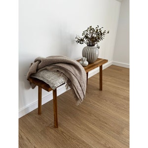 May include: A small, wooden bench with a rectangular top and four legs. A tan throw blanket and a patterned pillow rest on the bench. A vase with a striped design and a small white ceramic container sit on the bench. The bench is in a room with wood flooring.