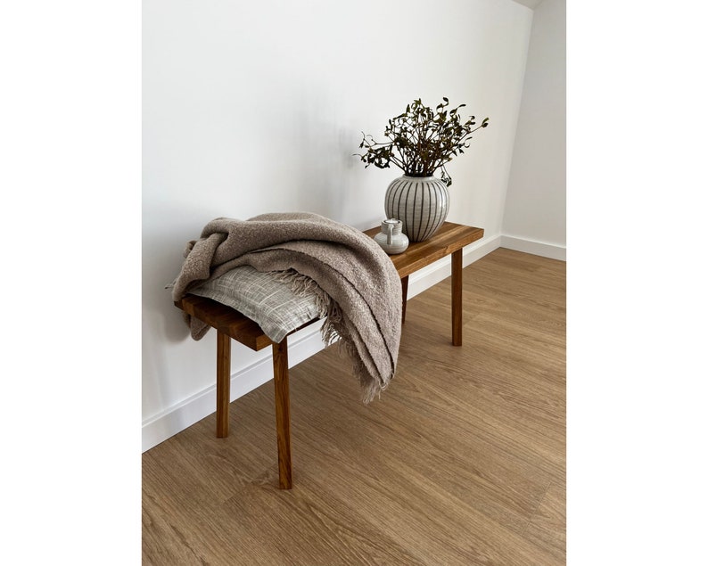 May include: A small, wooden bench with a rectangular top and four legs. A tan throw blanket and a patterned pillow rest on the bench. A vase with a striped design and a small white ceramic container sit on the bench. The bench is in a room with wood flooring.