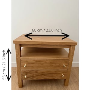 May include: A light brown wooden nightstand with an open shelf and two drawers featuring gold-toned knobs. The nightstand measures 23.6 inches in width and 21.6 inches in height, as indicated by the black arrows and text printed on the image.