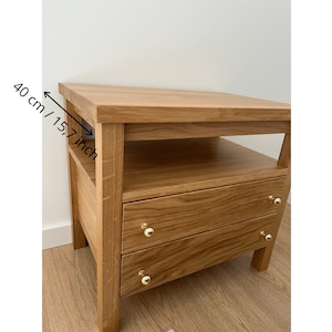 May include: A light brown wooden nightstand with two drawers and an open shelf. The nightstand features gold-toned round knobs on each drawer. A measurement indicator on the side shows a depth of 15.7 inches for the top surface.