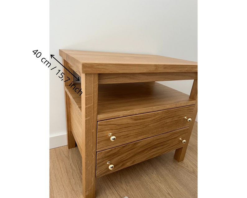 May include: A light brown wooden nightstand with two drawers and an open shelf. The nightstand features gold-toned round knobs on each drawer. A measurement indicator on the side shows a depth of 15.7 inches for the top surface.