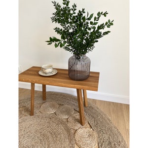 Puede incluir: Una mesa de madera con un acabado natural y tres patas se encuentra sobre una alfombra tejida. Un jarrón con una planta verde frondosa se encuentra sobre la mesa.