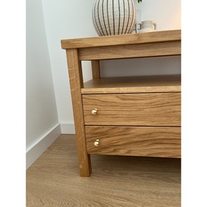 May include: A light brown oak wood console table featuring two horizontal drawers with small, round, polished gold-tone metal knobs. The piece has a rectangular top surface and an open shelf located directly beneath the main tabletop.