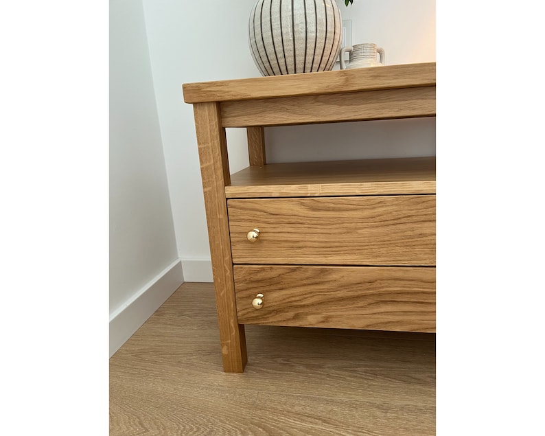 May include: A light brown oak wood console table featuring two horizontal drawers with small, round, polished gold-tone metal knobs. The piece has a rectangular top surface and an open shelf located directly beneath the main tabletop.