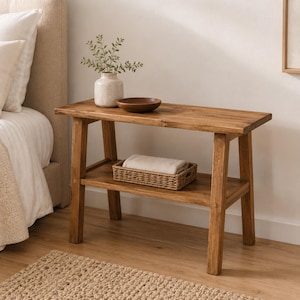 May include: A rectangular wooden console table with a natural finish, featuring four angled legs and a lower shelf. On the top surface sits a white ceramic vase with greenery and a small wooden bowl. A woven basket with a folded cloth rests below.
