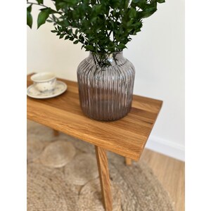 May include: A translucent, smoky grey glass vase with a vertical ribbed texture. The vase has a wide, rounded body and a narrow neck, containing a cluster of dark green leafy branches. The glass reflects light along its ridges.