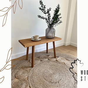 May include: A rectangular wooden bench with four straight legs, featuring a light brown wood grain finish. On the bench sits a ceramic cup and saucer with a speckled pattern, alongside a ribbed glass vase containing green leafy branches.