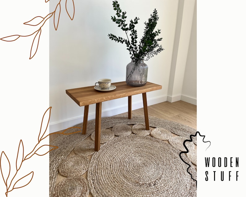 May include: A rectangular wooden bench with four straight legs, featuring a light brown wood grain finish. On the bench sits a ceramic cup and saucer with a speckled pattern, alongside a ribbed glass vase containing green leafy branches.