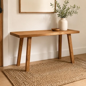 May include: A rectangular wooden console table with four angled legs, featuring a natural light brown wood grain finish. On the table sits a matte off-white ceramic vase containing green leafy branches and a small, round, dark brown wooden bowl.