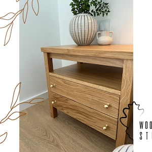 May include: A light oak nightstand featuring an open shelf and two drawers with small, round, gold-toned metal knobs. The wooden furniture has a visible grain pattern and is topped with a striped ceramic vase containing green foliage.