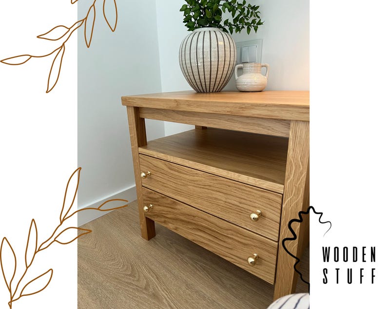 May include: A light oak nightstand featuring an open shelf and two drawers with small, round, gold-toned metal knobs. The wooden furniture has a visible grain pattern and is topped with a striped ceramic vase containing green foliage.