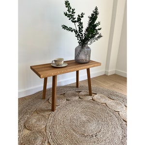 May include: A rectangular wooden bench with four straight legs, featuring a natural wood grain finish. On the bench sits a small ceramic cup and saucer with a speckled pattern, alongside a ribbed glass vase containing green leafy branches.