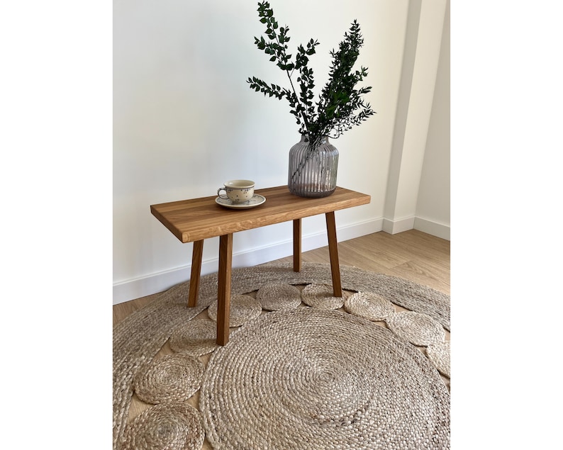 May include: A rectangular wooden bench with four straight legs, featuring a natural wood grain finish. On the bench sits a small ceramic cup and saucer with a speckled pattern, alongside a ribbed glass vase containing green leafy branches.