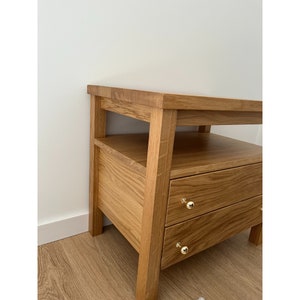 May include: A light brown oak wood nightstand featuring an open shelf and two drawers. Each drawer is equipped with a small, round, polished gold-tone metal knob. The piece has a rectangular top surface and four sturdy legs, showcasing a natural wood grain.