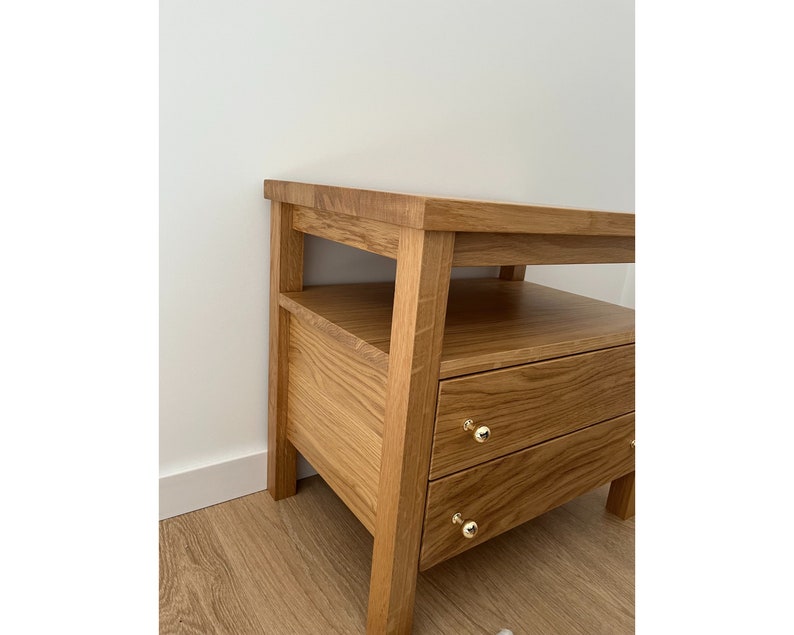 May include: A light brown oak wood nightstand featuring an open shelf and two drawers. Each drawer is equipped with a small, round, polished gold-tone metal knob. The piece has a rectangular top surface and four sturdy legs, showcasing a natural wood grain.