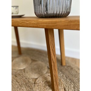 May include: A light brown wooden bench with four straight, tapered legs and a smooth, rectangular top surface. The wood grain is visible throughout the structure, showcasing a natural finish. A ribbed, translucent gray glass vase sits on top.