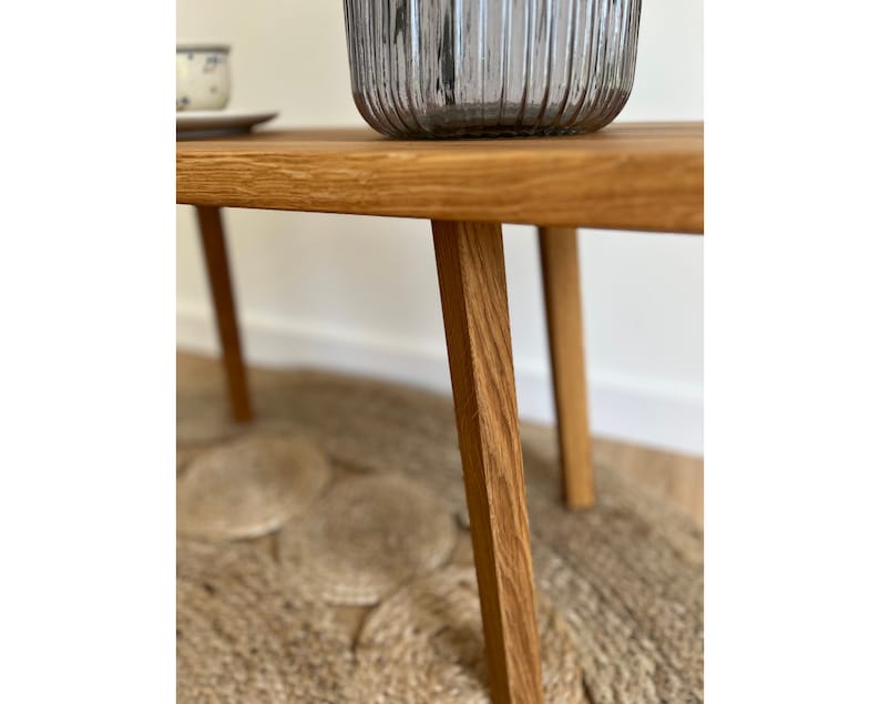 May include: A light brown wooden bench with four straight, tapered legs and a smooth, rectangular top surface. The wood grain is visible throughout the structure, showcasing a natural finish. A ribbed, translucent gray glass vase sits on top.