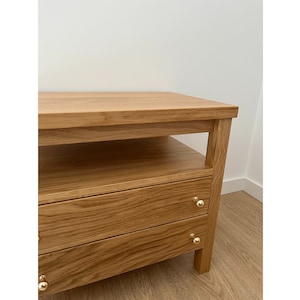 May include: A light brown wooden nightstand featuring a flat top surface, an open storage shelf below the top, and two drawers with small, round, polished gold-colored metal knobs. The wood grain is visible across the entire structure.