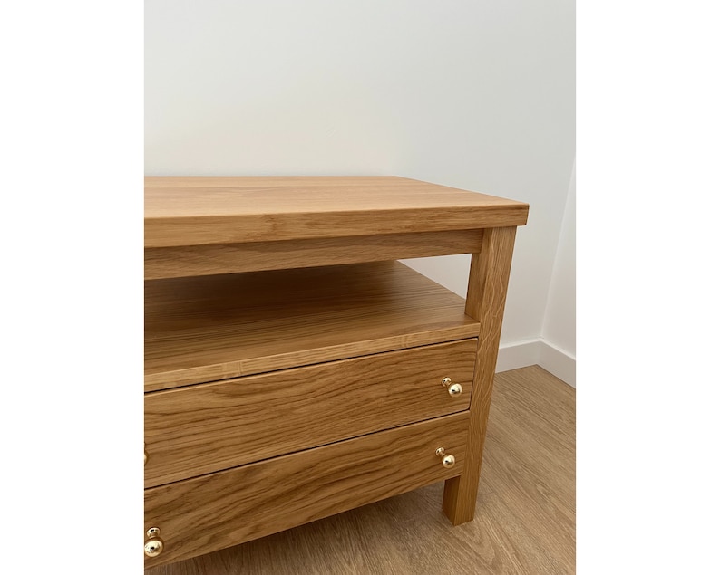 May include: A light brown wooden nightstand featuring a flat top surface, an open storage shelf below the top, and two drawers with small, round, polished gold-colored metal knobs. The wood grain is visible across the entire structure.