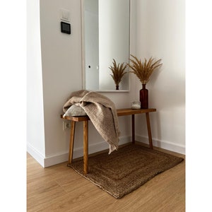 May include: A cozy entryway scene featuring a wooden bench with a tan throw blanket and a decorative pillow. A brown jute rug sits beneath the bench. A brown glass vase with dried plants and a small candle add to the warm, inviting atmosphere.