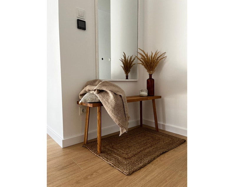 May include: A cozy entryway scene featuring a wooden bench with a tan throw blanket and a decorative pillow. A brown jute rug sits beneath the bench. A brown glass vase with dried plants and a small candle add to the warm, inviting atmosphere.