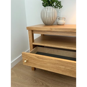 May include: A light oak wood media console featuring a single open shelf and one pull-out drawer with a small, round, polished gold-tone knob. The drawer interior is lined with a dark charcoal material, and the unit has a rectangular frame.