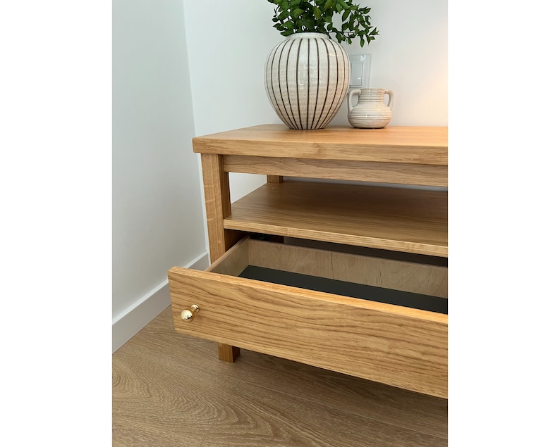 May include: A light oak wood media console featuring a single open shelf and one pull-out drawer with a small, round, polished gold-tone knob. The drawer interior is lined with a dark charcoal material, and the unit has a rectangular frame.