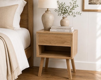 May include: A light oak nightstand with one drawer and an open shelf, supported by four angled legs. On top sits a beige ceramic table lamp with a white shade, a small ceramic vase with green foliage, and two books titled The Kinfolk Home and This Is Home.