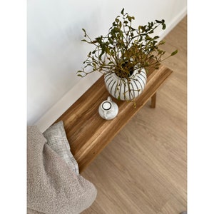 May include: A wooden bench with a white and black striped vase holding greenery. A small white ceramic container sits on the bench. A beige throw and a gray pillow are on the bench. The bench is on a wood floor.