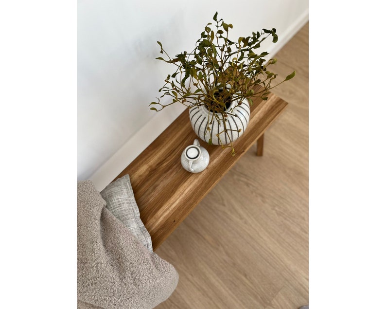 May include: A wooden bench with a white and black striped vase holding greenery. A small white ceramic container sits on the bench. A beige throw and a gray pillow are on the bench. The bench is on a wood floor.