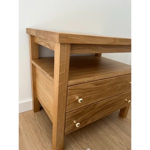 May include: A light brown wooden nightstand featuring a flat top surface, an open shelf, and two drawers. Each drawer is equipped with a small, round, polished gold-tone metal knob. The piece is constructed from wood with a visible natural grain pattern.
