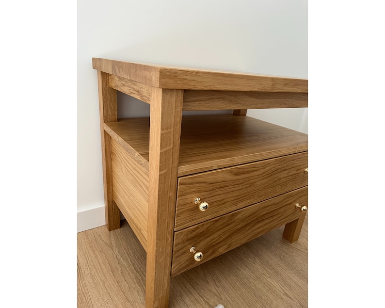 May include: A light brown wooden nightstand featuring a flat top surface, an open shelf, and two drawers. Each drawer is equipped with a small, round, polished gold-tone metal knob. The piece is constructed from wood with a visible natural grain pattern.
