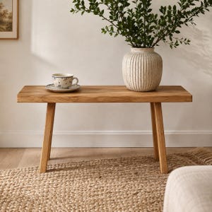 May include: A rectangular wooden bench with four angled legs. On the bench sits a white ceramic teacup with a blue floral pattern on a matching saucer, and a large, ribbed, off-white ceramic vase containing a leafy green plant.