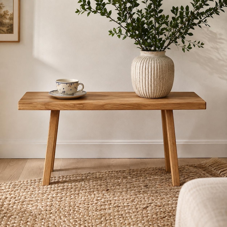 May include: A rectangular wooden bench with four angled legs. On the bench sits a white ceramic teacup with a blue floral pattern on a matching saucer, and a large, ribbed, off-white ceramic vase containing a leafy green plant.