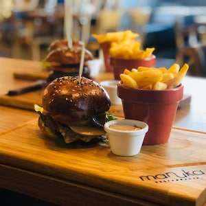 Puede incluir: Una tabla de cortar de madera con una hamburguesa, patatas fritas y un pequeño cuenco de salsa. La tabla de cortar tiene la palabra "manuka" impresa.