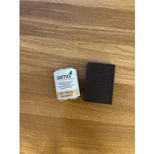 May include: A small metal can of Osmo 1101 Farblos Transparent wood finish and a dark brown sanding block sit on a light brown wooden surface. The can has a white label with green and black text.