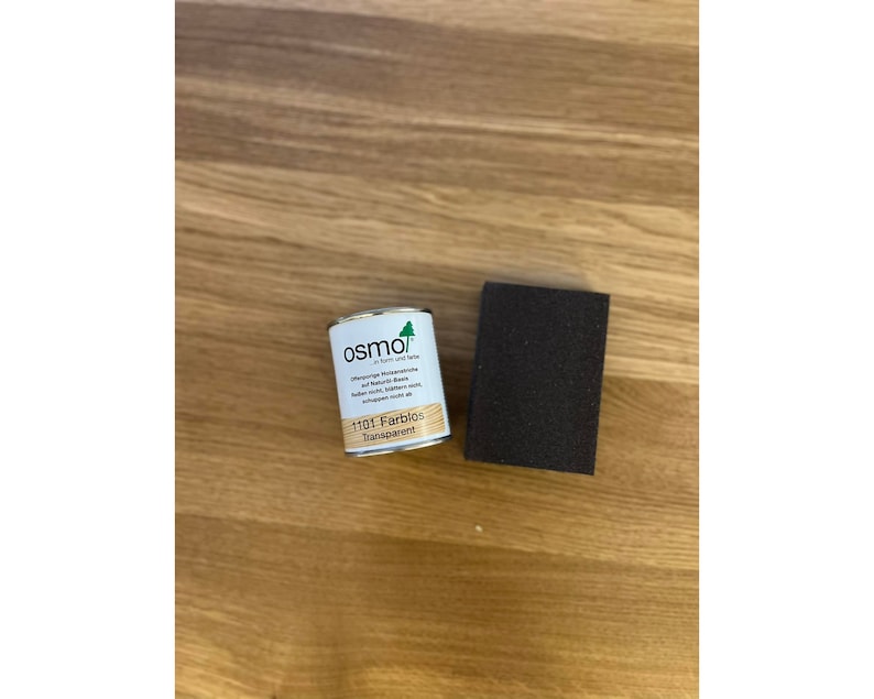 May include: A small metal can of Osmo 1101 Farblos Transparent wood finish and a dark brown sanding block sit on a light brown wooden surface. The can has a white label with green and black text.