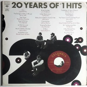Op de afbeelding: Een zwart-witte vinyl platenhoes met de titel "20 Years of #1 Hits" en een lijst met nummers en artiesten. De platenhoes heeft een groot nummer "20" in zwart-wit met een rood Columbia-logo in het midden.