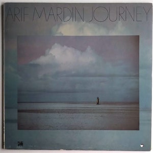 Puede incluir: Una portada de álbum de vinilo vintage con una fotografía de una figura solitaria de pie en una playa con un cielo nublado en el fondo. El título del álbum "ARIF MARDIN JOURNEY" está impreso en letras negras en la parte superior de la portada.