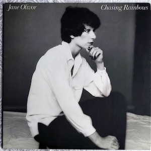 May include: Black and white photograph of a woman with short dark hair, wearing a white button-down shirt and black trousers. The photo is from a record album cover and includes the text "Jane Olivor" and "Chasing Rainbows".