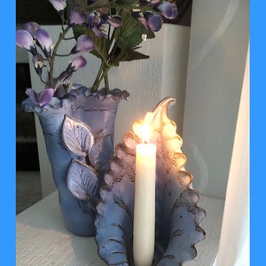 May include: A blue ceramic vase with a ruffled top and leaf design holds a bouquet of purple flowers. Next to it is a blue ceramic candle holder shaped like a leaf with a white candle inside.