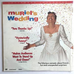 May include: A laserdisc cover for the movie "Muriel's Wedding". The cover features a woman in a white wedding dress with confetti falling around her. The text on the cover reads "Muriel's Wedding" and includes quotes from critics, such as "Two Thumbs Up!" and "Hysterically Funny!"